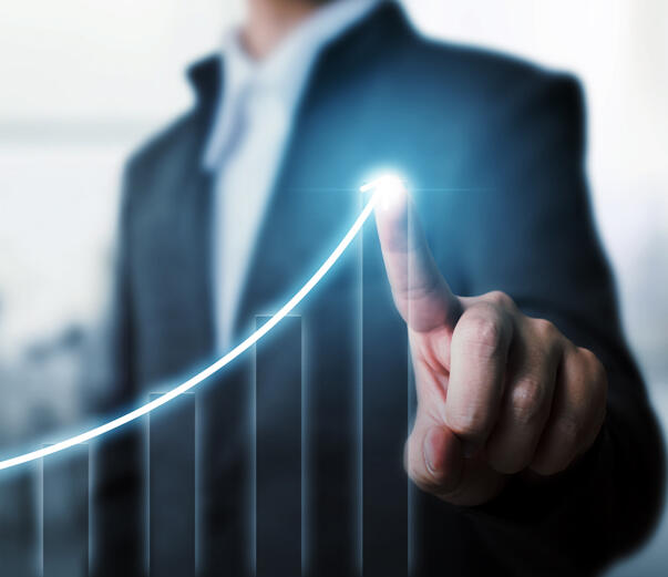 Emeriti Consulting - Grow The image depicts a business professional in a blurred background, pointing to the peak of a glowing upward-trending graph. The graph symbolizes growth, success, or progress, and the bright light at the fingertip emphasizes achievement or a key milestone.