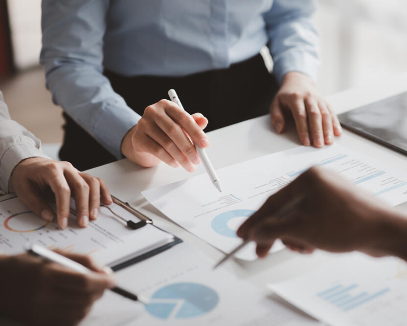 Emeriti Consulting - Learn The image shows a group of professionals collaborating around a table. Their hands are visible, engaged in discussion and pointing at documents with charts and graphs. A pen and tablet are also on the table, suggesting a focus on data analysis or project p
