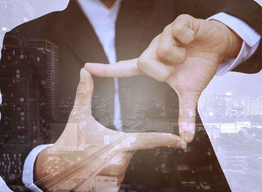 Emeriti Consulting - Thrive The image shows a close-up of a man wearing a brown suit, forming a frame shape with his hands in front of his chest.