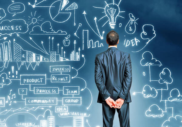 Emeriti Consulting - Plan The image depicts a man in a formal suit standing with his back to the viewer, observing a large, illuminated wall filled with intricate diagrams, charts, and illustrations. The visuals represent concepts like success, ideas, teamwork, and processes.